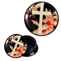 Load image into Gallery viewer, An open Schraub Plug Acryl Piercing in black with a white cross and pink hibiscus flowers on a dark background. The container, labeled &quot;rosa beige weiß Kreuz vor Blumen,&quot; is round and shows its empty interior.
