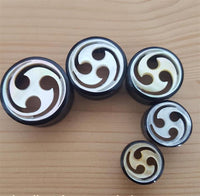 Load image into Gallery viewer, Five round black Horn Plug Tunnel Perlmutt Tomoe Inlay weiß double flared, featuring elegant white swirl inlays and arranged by size from largest to smallest on a light wooden surface—ideal for tribal piercing fans.
