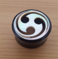Load image into Gallery viewer, The Horn Plug Tunnel Perlmutt Tomoe Inlay weiß double flared features a glossy white tomoe inlay on round black horn, stylishly set on light wood—ideal for fans of distinctive tribal piercing designs.
