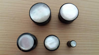 Load image into Gallery viewer, Five Buffalo Horn Plug Perlmutt Inlay weiß handgeschnitzt Lobe Piercing Tunnels, arranged on a light wooden surface, feature shiny white tops and differ in size, with one tunnel significantly smaller than the rest.
