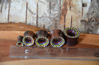 Load image into Gallery viewer, Five Holz Tunnel Sonoholz handbemalt bunt Punkte Plug Polka Dots wooden ear plugs, each larger than the last and featuring colorful polka dot designs, are displayed with a clear ruler on rough wood.
