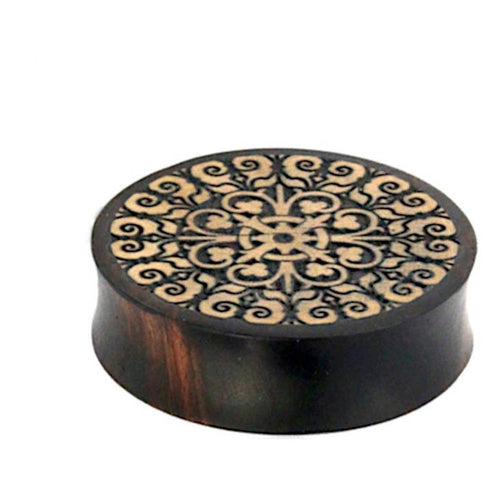 A round, dark wooden box with a flat lid displays an intricate symmetrical cross and scrollwork pattern in light wood, echoing the hand-carved style of the Holz Flesh Plug Tamarinde Holz Krokodilholz Kreuz Ranken.