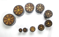 Cargar imagen en el visor de la galería, Nine round black Plug Arangholz Wikinger Kompass Inlay double flared plugs in various sizes are arranged in a curve on a white background, each with a gold Viking rune symbol for a unique and bold set.
