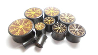 Cargar imagen en el visor de la galería, A set of Plug Arangholz Wikinger Kompass Inlay double flared plugs in black, each with a golden Icelandic Vegvisir symbol on the front, displayed on a white background—ideal for fans of unique Holzplug style and Arangholz craftsmanship.
