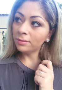 Load image into Gallery viewer, A woman with long, light brown hair looks to the side, wearing a grey top and Ohstecker Shiva Auge rund 925 Silber earrings. She has a ring on her left hand near her collar against a softly blurred outdoor background.
