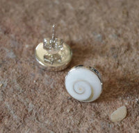Load image into Gallery viewer, A pair of Ohstecker Shiva Auge rund 925 Silber (6mm, 10mm, 12mm) with white spiral shell inlays is shown resting on a textured brown stone surface.
