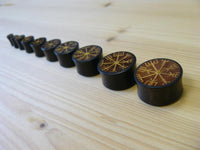 Cargar imagen en el visor de la galería, A row of Plug Arangholz Wikinger Kompass Inlay double flared plugs, engraved with yellow Norse runes including a Vegvisir, are arranged from largest to smallest on a light wooden surface.
