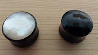 Load image into Gallery viewer, Two Buffalo Horn Plug Perlmutt Inlay weiß handgeschnitzt Lobe Piercing Tunnels rest on wood: the left with a shiny white Perlmutt inlay, the right solid black—ideal organic expanders for stretched piercings.
