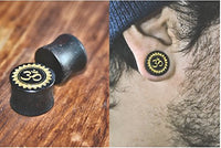 Cargar imagen en el visor de la galería, A close-up of an ear wearing a black gauge with a gold Om symbol. Next to it, two matching Holz Plug Tamarind Wood Blume Om gold, hand-carved and laser-engraved tribal tunnels, are displayed on wood.
