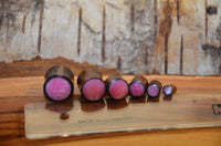Load image into Gallery viewer, Six Holz Plug Sonoholz rosa schillernd Resin Inlay double flared plugs in graduated sizes, with pink centers, are lined up beside a transparent ruler on a wood-textured surface.
