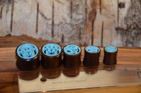 Load image into Gallery viewer, Five Tunnel Sonoholz Resin Türkis Inlay Spiralbögen, each with carved rose designs, are arranged by size beside a transparent ruler on a wooden surface, with a textured bark backdrop.
