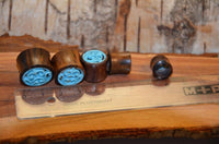 Load image into Gallery viewer, The Tunnel Sonoholz Resin Türkis Inlay Spiralbögen, along with two other wooden plugs, are arranged beside a clear ruler on a rustic wooden surface.

