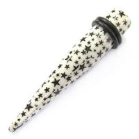 Load image into Gallery viewer, The Sterne Piercing Expander Dehnungsstab is a white, straight unisex ear stretching stick with black star patterns in various sizes and a black rubber O-ring near the wider end.
